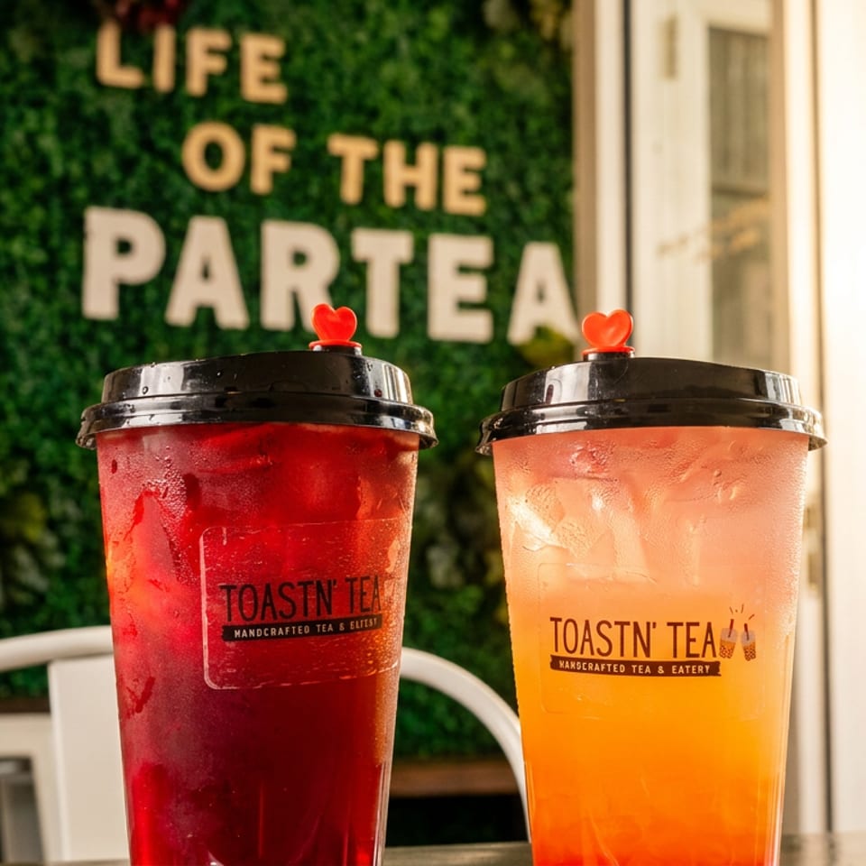 Two Toastn' Tea drinks, one red and one orange, on a table with a "Life of the Partea" sign.