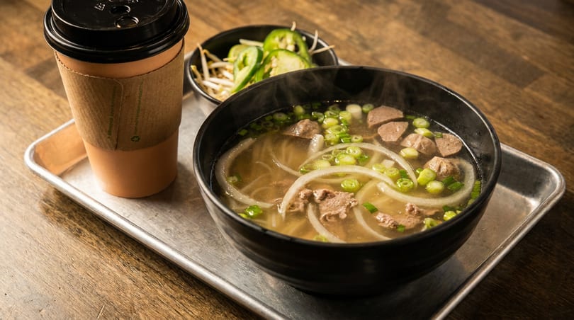 Steaming bowl of pho with beef, onions, and scallions, alongside a coffee and side of sprouts.