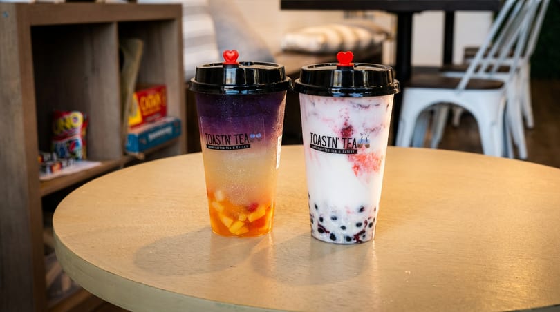 Two Toastn Tea bubble tea drinks, one purple and orange, one white and red, on a wooden table.