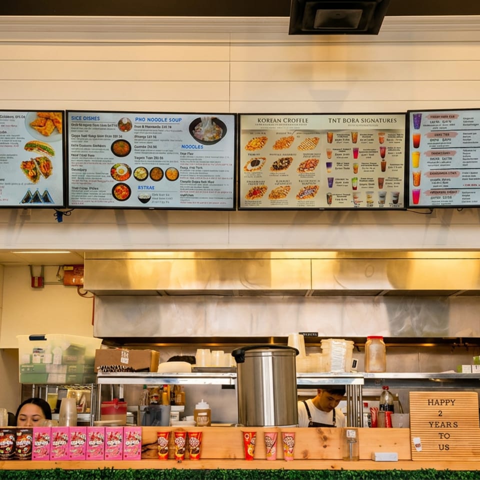 Digital menu boards displaying food and drink options above a busy restaurant kitchen counter.