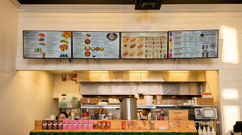 Digital menu boards displaying food and drink options above a busy restaurant kitchen counter.
