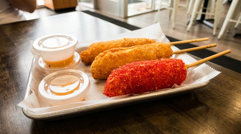 Three Korean corn dogs, one red and two golden, with two dipping sauces on a metal tray.