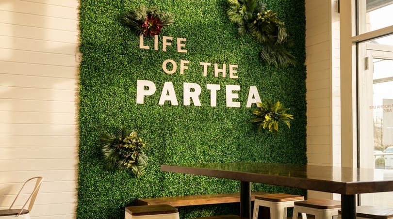 Green faux-grass wall with "LIFE OF THE PARTEA" in wooden letters, next to a table and benches.