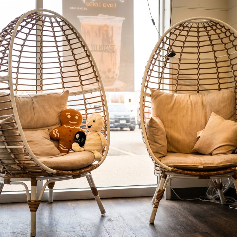 Two wicker egg chairs with pillows, one holding stuffed toys, by a window with a drink ad.