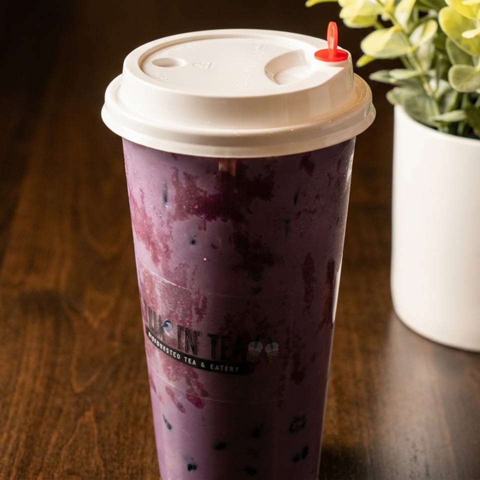 A purple bubble tea with a white lid and red stopper sits on a wooden table next to a plant.