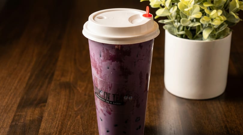 A purple bubble tea with a white lid and red stopper sits on a wooden table next to a plant.
