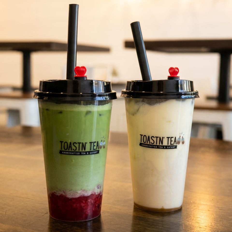Two Toastn' Tea drinks, one green with red fruit and one white, on a wooden table.