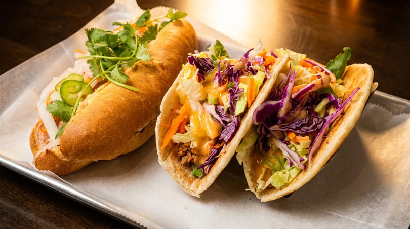 A banh mi sandwich and two tacos with colorful toppings on a metal tray with parchment paper.