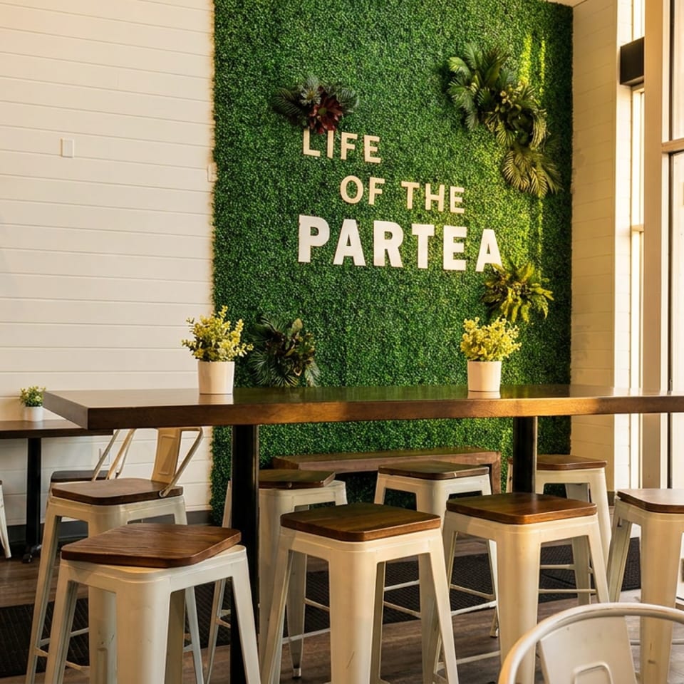 Modern cafe interior with a green moss wall featuring "LIFE OF THE PARTEA" text, tables, and stools.