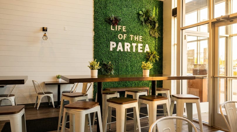 Modern cafe interior with a green moss wall featuring "LIFE OF THE PARTEA" text, tables, and stools.