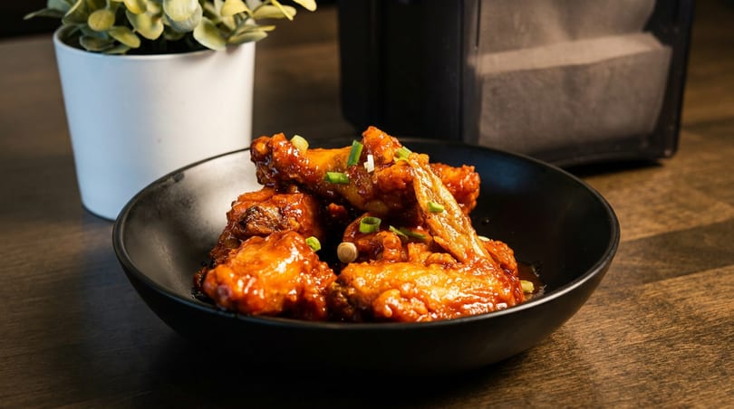 A black bowl filled with saucy chicken wings topped with chopped green onions on a wooden table.