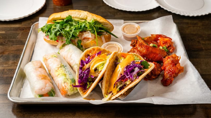 A tray of Vietnamese and Korean dishes, including a banh mi, spring rolls, tacos, and chicken wings.
