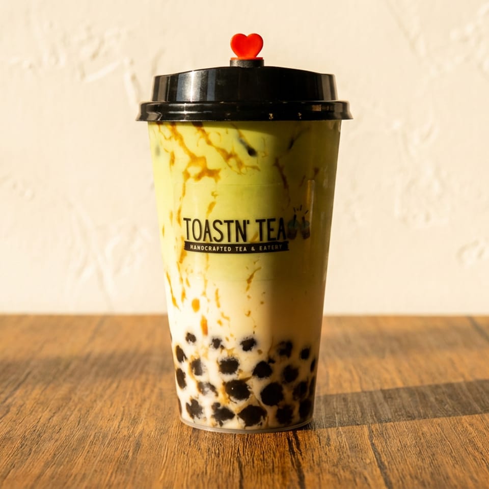 A Toastn' Tea boba drink with green tea and milk, topped with a red heart, on a wooden table.