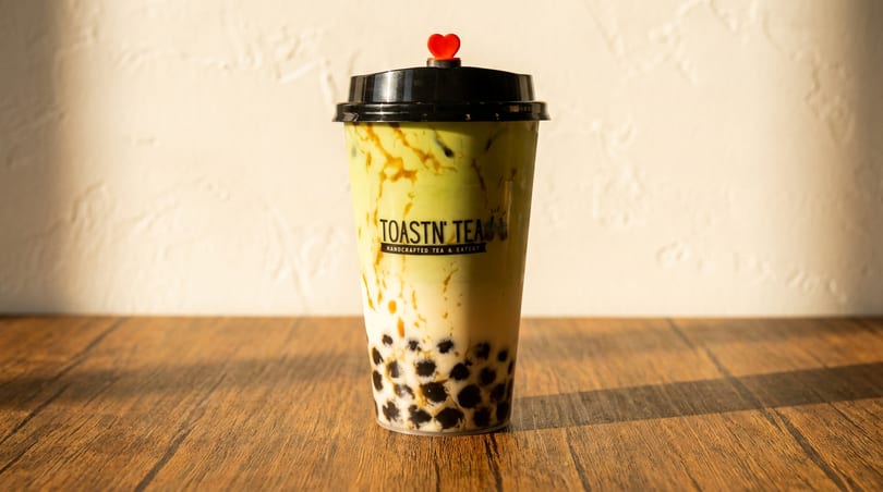 A Toastn' Tea boba drink with green tea and milk, topped with a red heart, on a wooden table.