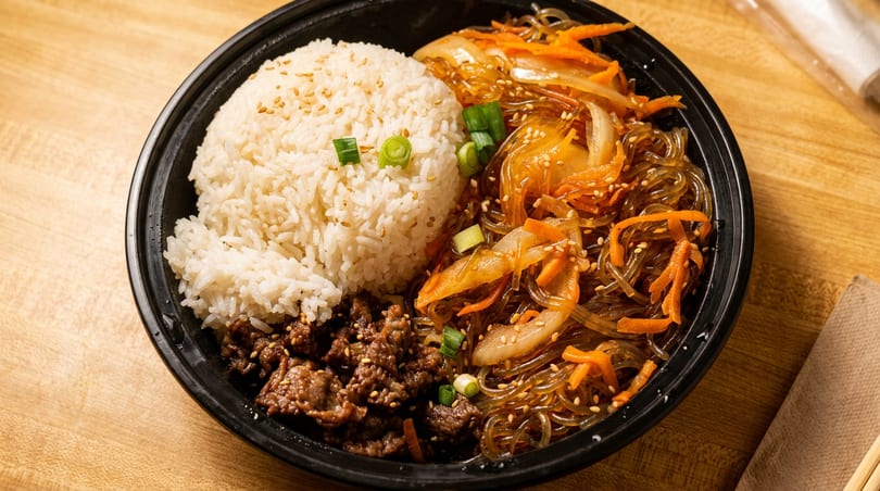 A black bowl filled with white rice, bulgogi beef, and japchae noodles with vegetables.