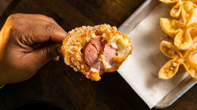 Hand holding a golden-brown corn dog, split open to reveal sausage and melted cheese inside.