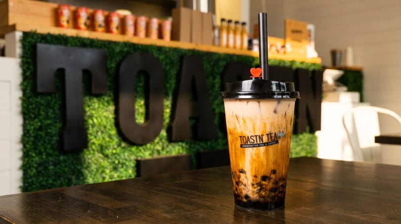 Toastn' Tea boba drink with a heart-shaped stopper on a wooden table, green wall in background.